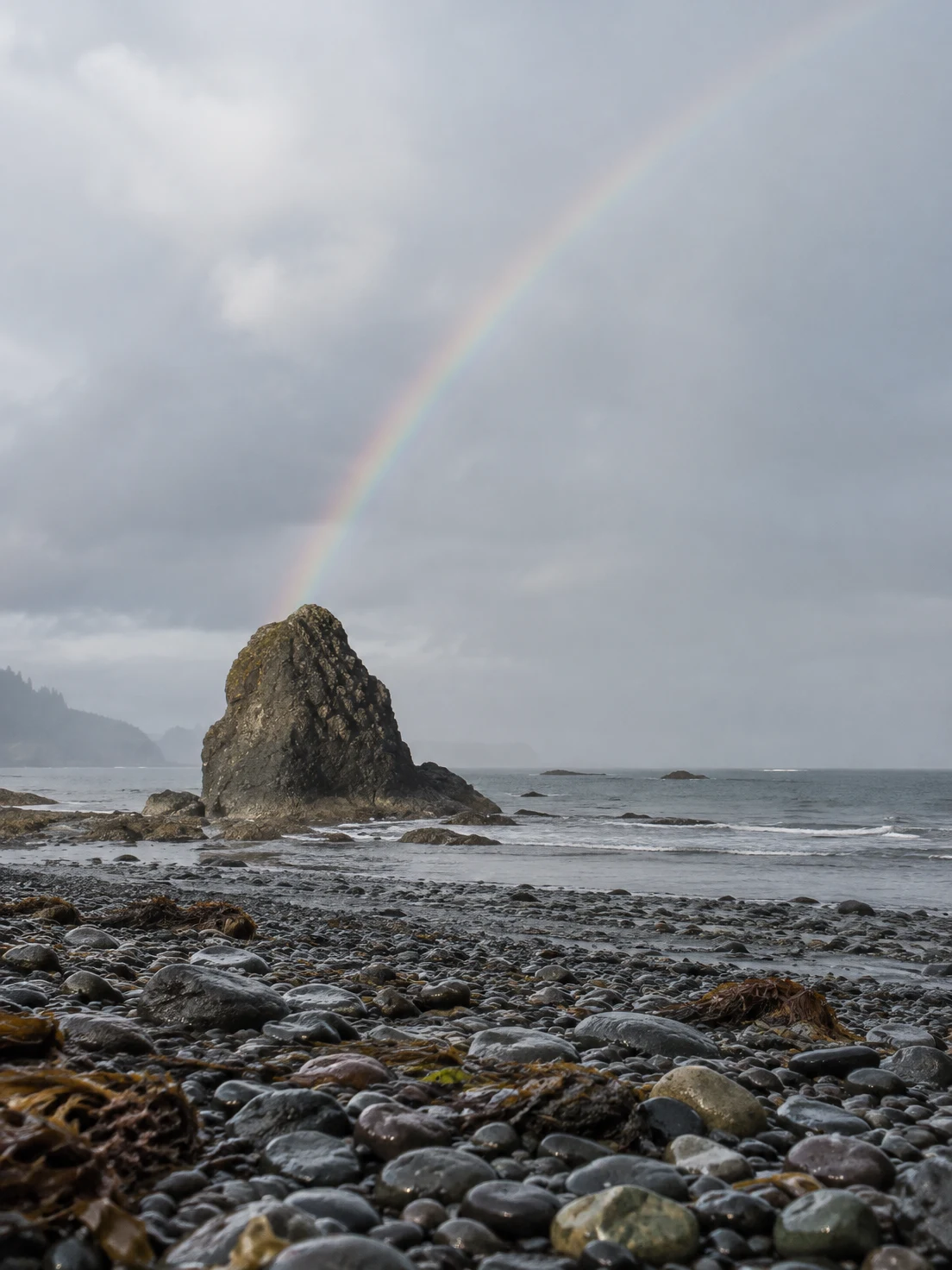 A dark sea stack stands on a wet shingle beach beneath a pale overcast sky, with a faint rainbow rising diagonally from its peak and grey sea behind it.