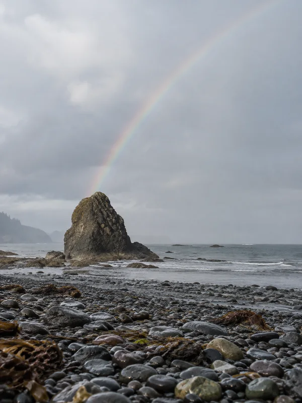 A dark sea stack stands on a wet shingle beach beneath a pale overcast sky, with a faint rainbow rising diagonally from its peak and grey sea behind it.