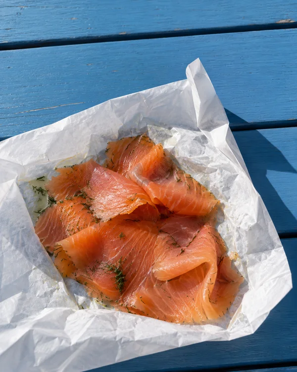 Overhead view of overlapping coral-pink salmon slices with dill flecks in an opened white paper parcel on a blue-painted wooden table, lit by direct sunlight…