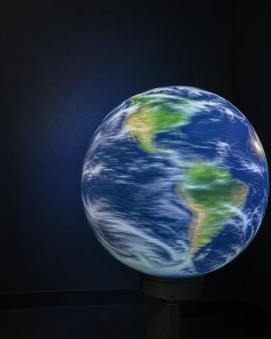 A luminous spherical Earth projection shows the Americas with soft lateral motion trails, mounted on a dark bracket against a charcoal wall with open dark…