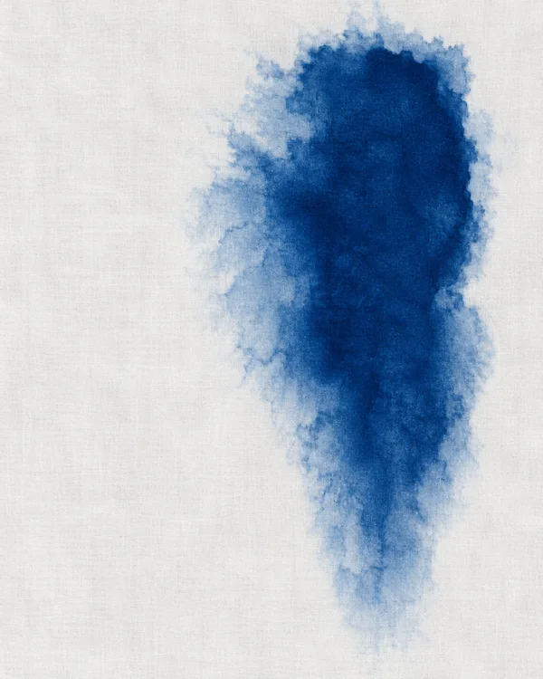 A tall irregular lapis-blue dye bloom with a soft, feathered pale-blue perimeter on chalk-white woven fabric, positioned right of center with a broad white…