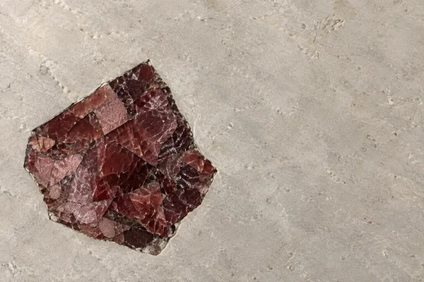 An irregular garnet-red crystalline patch with angular burgundy and dusty-rose facets lies left of center in a warm grey-beige stone surface, leaving broad…