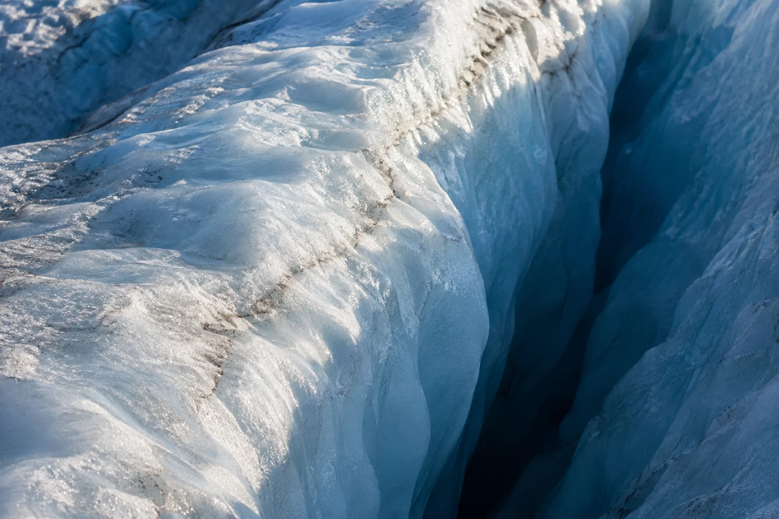 Close view of a glacier, with a sunlit pale blue ice ridge running diagonally beside a deep shadowed blue crevasse; small brown mineral streaks are visible in…