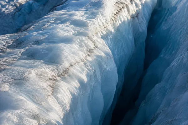 Close view of a glacier, with a sunlit pale blue ice ridge running diagonally beside a deep shadowed blue crevasse; small brown mineral streaks are visible in…