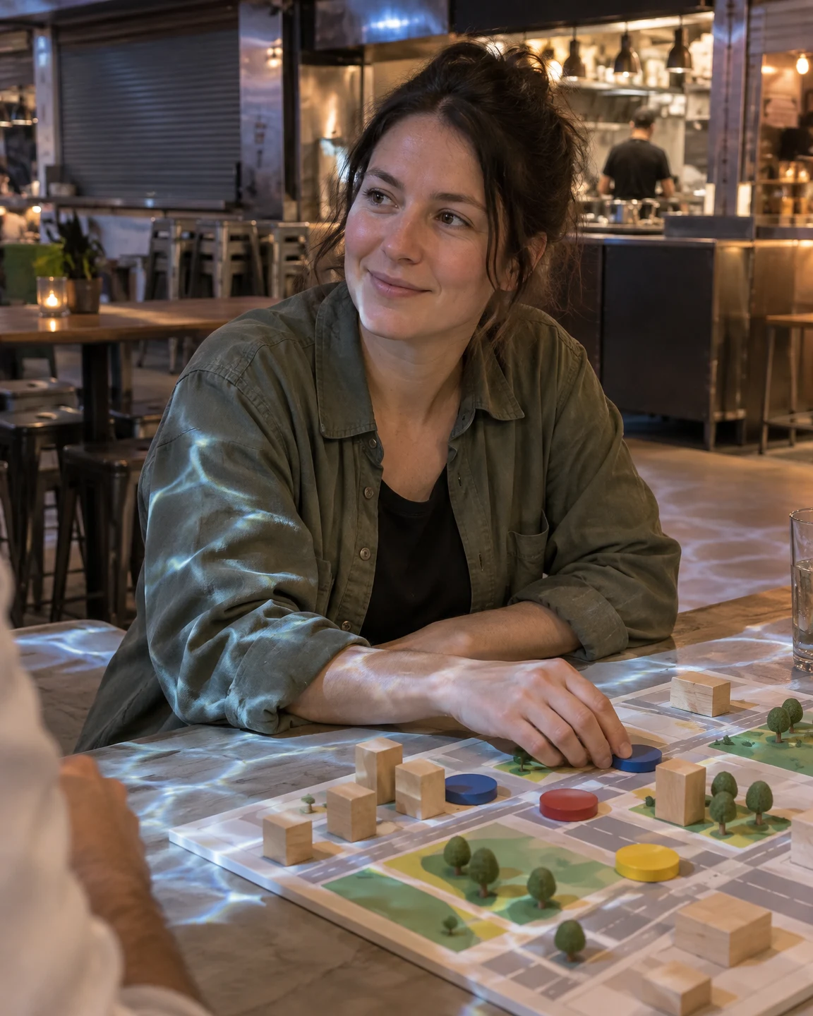 A woman in an olive overshirt sits at a communal table, smiling slightly as she looks off-frame.