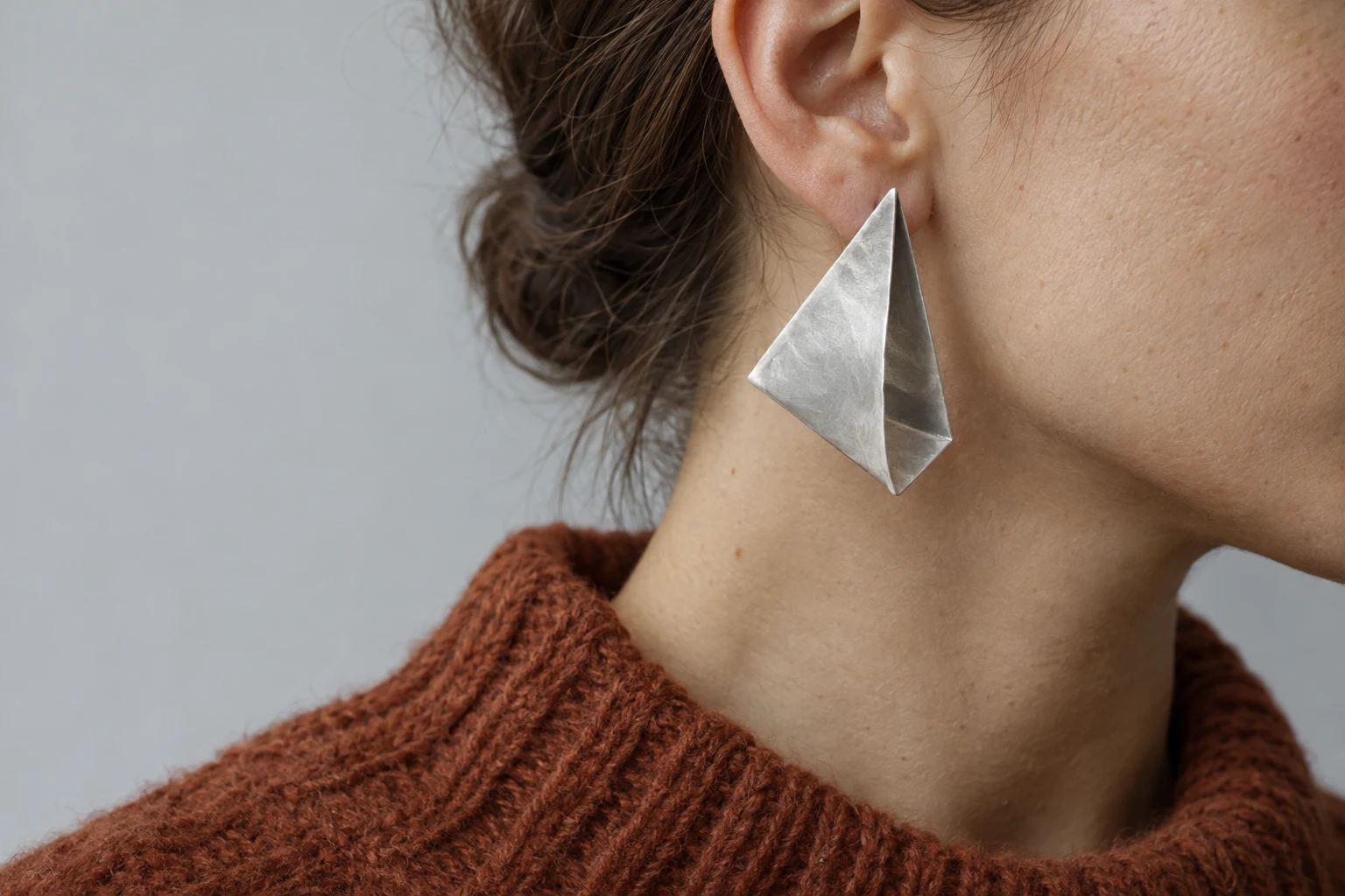 Tight side crop of an adult’s ear, jawline and neck with a large folded angular silver earring.
