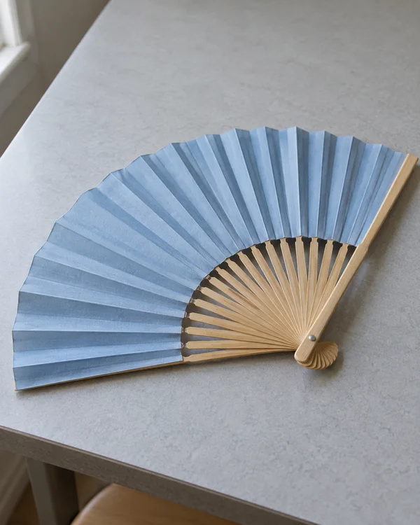 An open powder-blue paper fan with light bamboo ribs lies diagonally across a pale grey tabletop, with soft window light entering from the upper left and a…