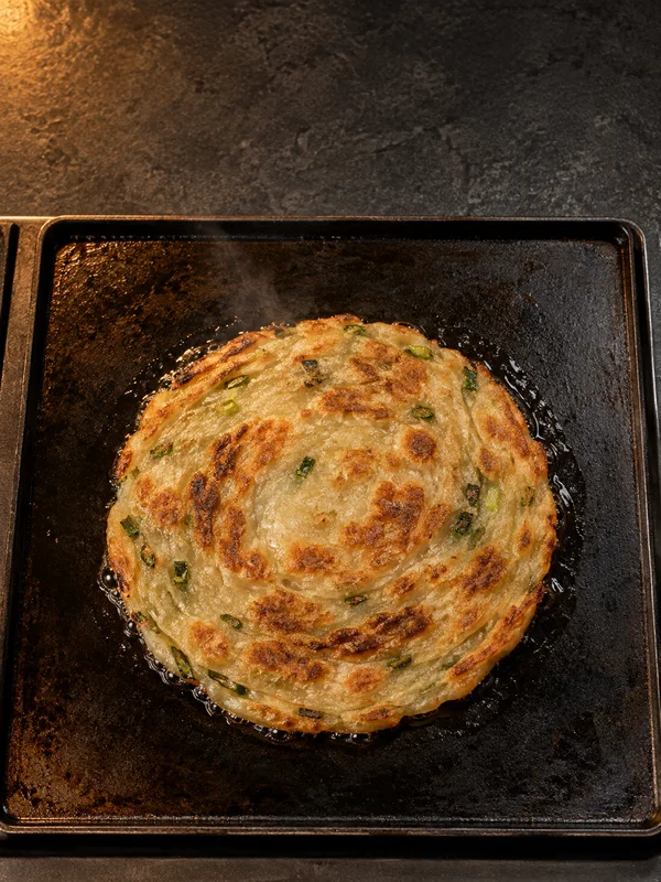 A golden scallion pancake with browned patches and green flecks cooks in oil on a dark square griddle set on a charcoal stone counter, viewed from overhead.