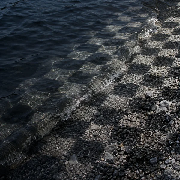 A dark lake wave curls diagonally onto a grey gravel shoreline, with scattered pale geometric reflections across the water and stones.