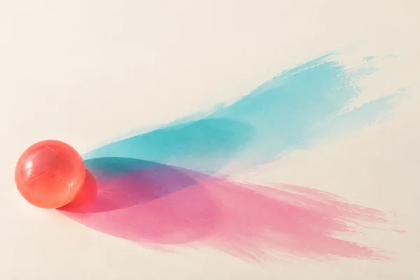 A translucent coral inflatable ball sits near the lower-left corner on a warm ivory surface, casting broad cyan and magenta brush-like shadows diagonally…