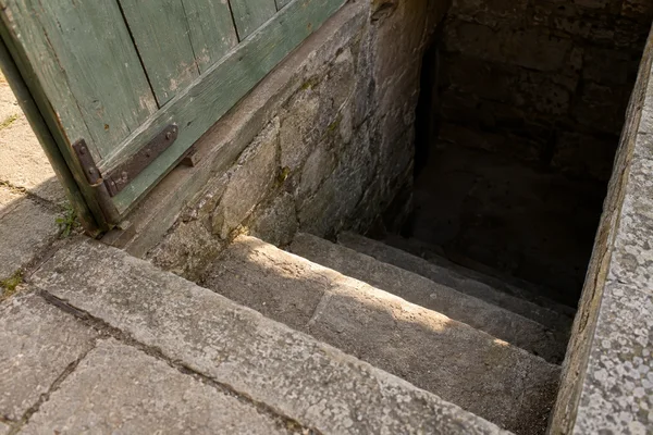 A faded green timber hatch stands open beside a rough stone cellar entrance, with worn steps descending into deep shadow and a band of sunlight crossing the…