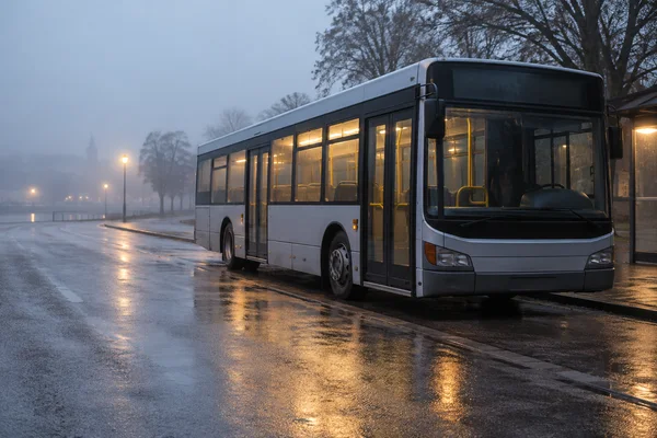 A silver city bus with amber-lit windows waits beside a wet curb on a foggy dawn road, with streetlamps, bare trees and open reflective pavement extending to…