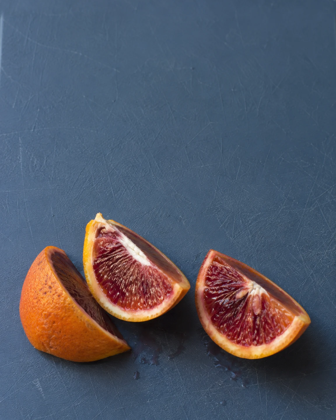 Three blood-orange wedges sit near the lower edge of a slate-blue cutting board: one is turned rind-side toward the camera, while two show their dark red cut…