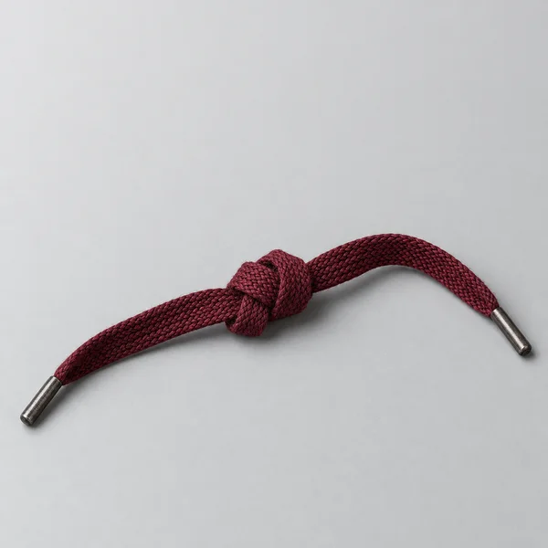 A dark burgundy braided lace is tied in a knot across a pale grey surface, with a metal tip at each end and ample empty space above.