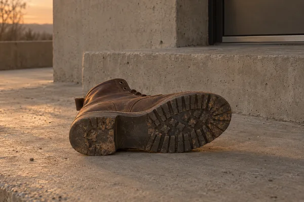 A worn walnut-brown leather work boot lies on its outer side on a concrete landing, its dusty rubber tread facing the camera; a raw-concrete step and dark…