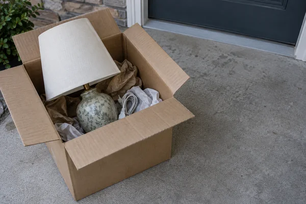 An open cardboard moving box sits on a concrete doorstep, holding a table lamp with an ivory shade, patterned ceramic base, crumpled packing paper and a coiled…