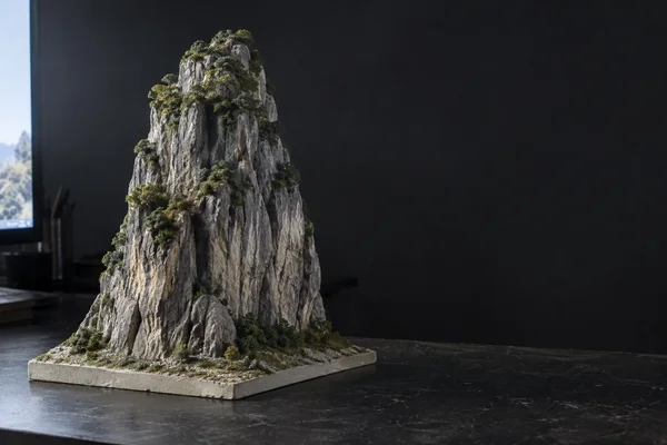 A detailed handmade model of a steep gray limestone tower with sparse green vegetation stands on a rectangular pale base atop a dark worktable.