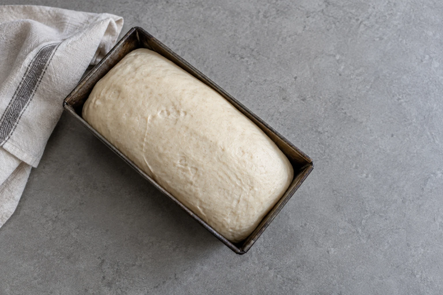 Overhead view of pale, risen bread dough in a well-used dark steel loaf tin on a cool-grey counter, with a folded off-white tea towel partly cropped beside it…