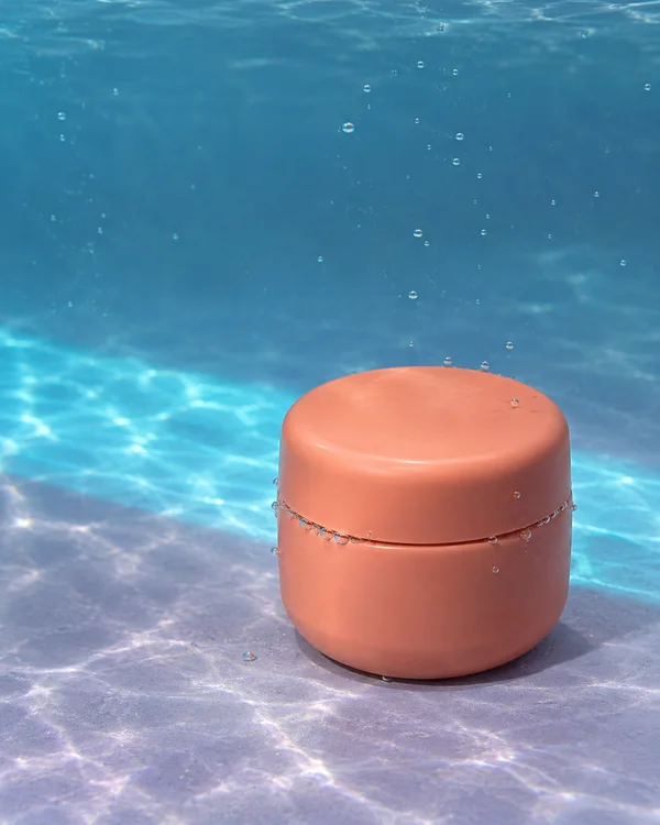 A blank coral-coloured cream jar with a closed screw lid sits on a pale lavender floor underwater.