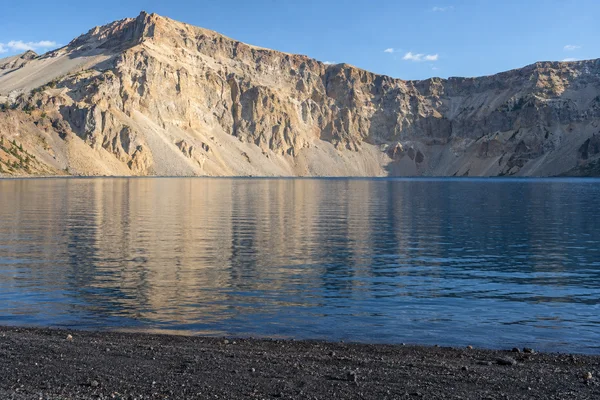 A deep-blue lake with faint ripples and broken reflections sits beneath a pale, rugged caldera wall; a dark gravel shoreline fills the foreground under a clear…