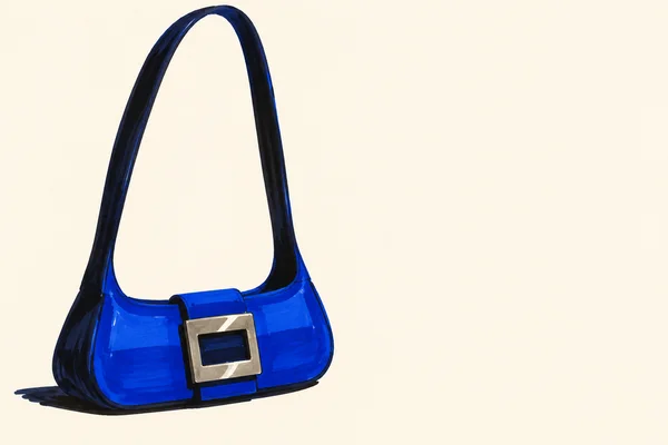 A cobalt-blue handbag with a very tall loop strap and a large silver rectangular buckle sits against a warm ivory background, with a dark marker shadow beneath…