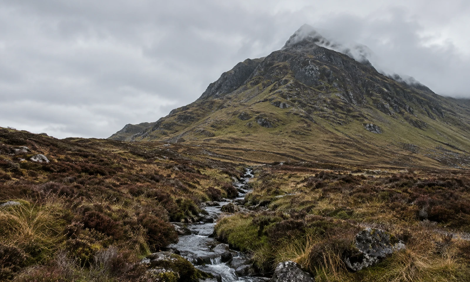 A narrow rocky stream winds through wet heather and tussock grass toward a steep, dark mountain whose summit is partly wrapped in low grey cloud beneath an…