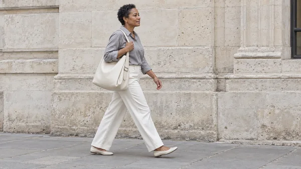 A woman with short natural hair walks along a stone facade in a silver-grey shirt, off-white trousers and pale flats, carrying a large ivory shoulder bag.
