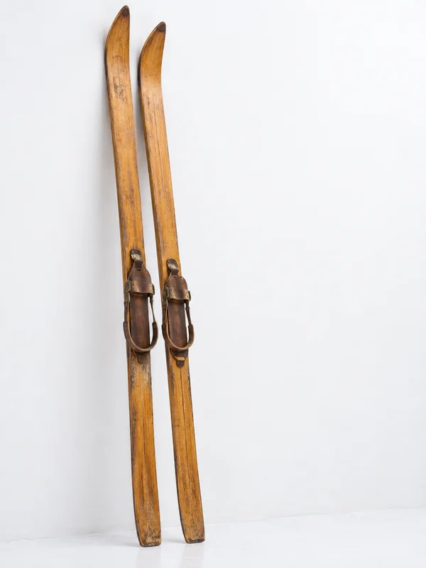 A matched pair of aged wooden skis with leather bindings stands against a bright white wall, with the skis left of center and open space to the right.