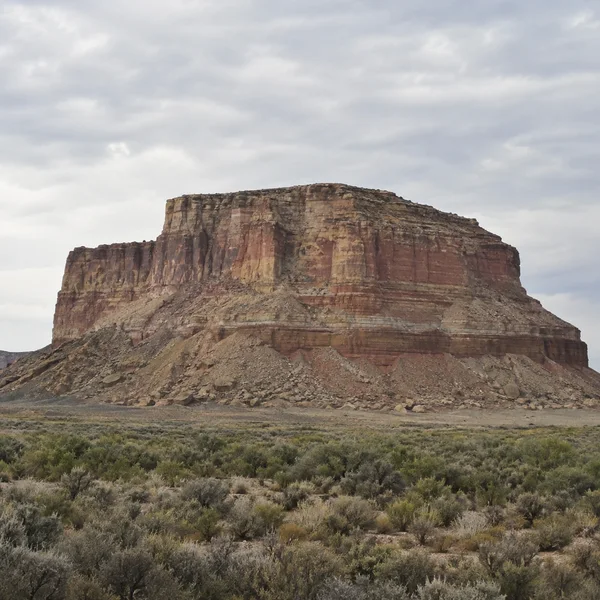A broad, flat-topped sandstone mesa with rust, ochre and grey horizontal bands rises above a rocky talus slope and a foreground plain of sagebrush beneath a…