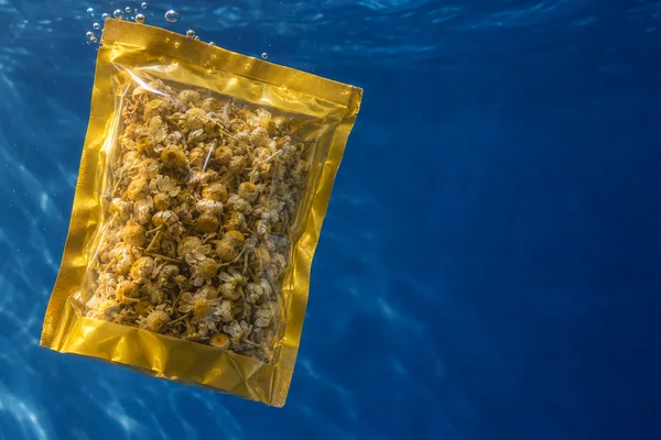 A clear, yellow-bordered rectangular pouch packed with dried chamomile flowers floats diagonally on the left against a deep blue underwater background, with…