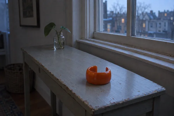 A glossy persimmon-orange open cuff rests near the front of a worn pale-grey wooden console beneath a window.
