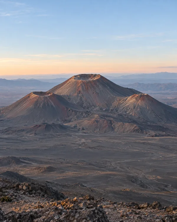A dormant volcanic massif with a large central crater cone and two smaller crater cones rises above a broad ash-grey plateau at dawn, with distant mountain…