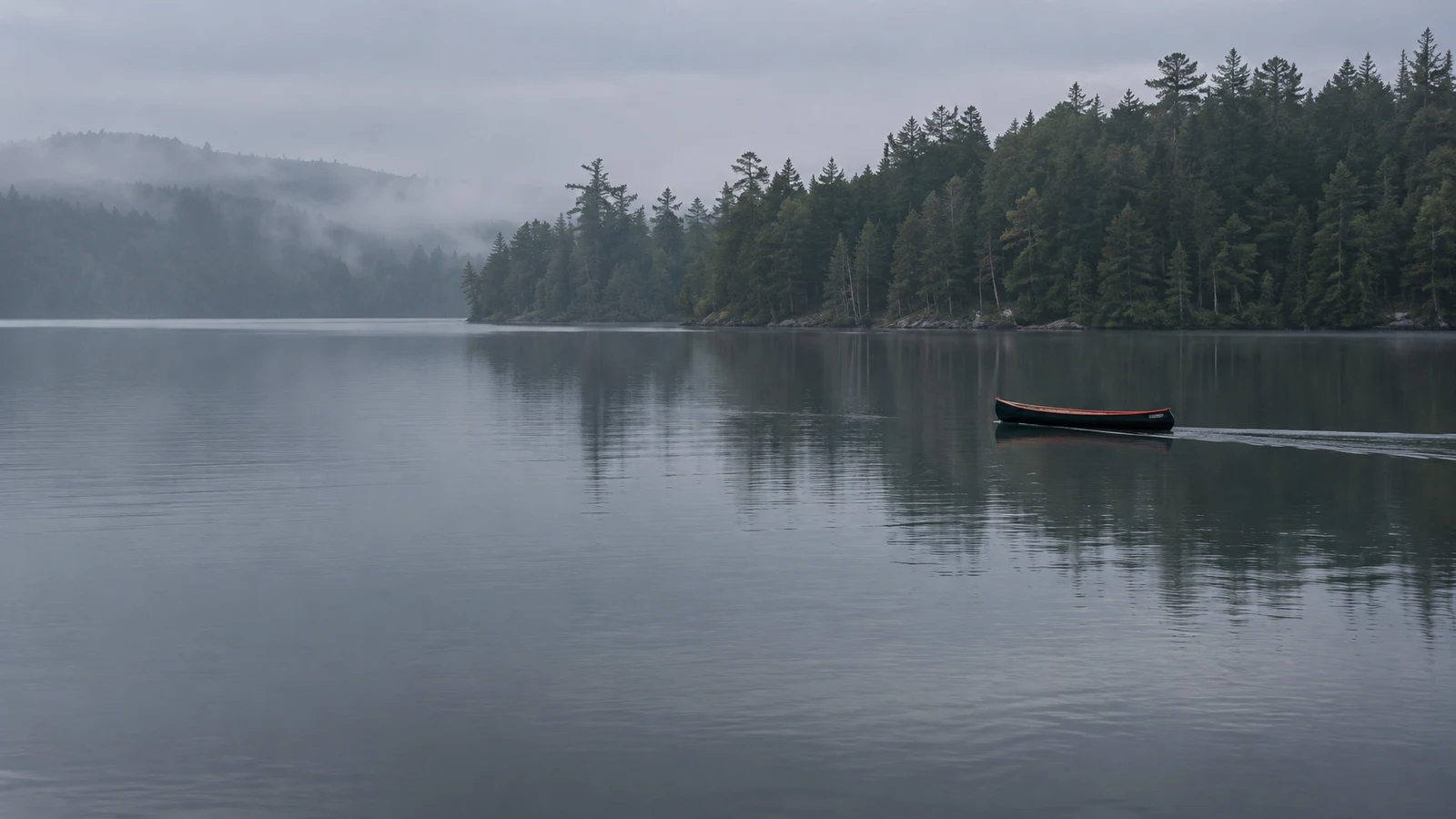 A slim black canoe with a coral-colored rim floats on a calm dark lake, with a misty evergreen shoreline behind it and broad open water in the foreground.