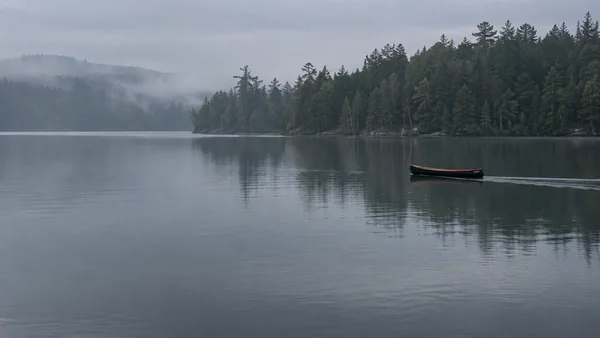 A slim black canoe with a coral-colored rim floats on a calm dark lake, with a misty evergreen shoreline behind it and broad open water in the foreground.