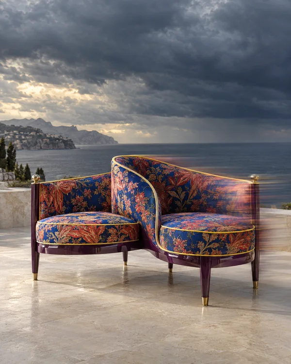 A sculptural two-seat bench with a curved central back, blue and coral floral upholstery, yellow piping, plum-colored frame and brass accents sits on a pale…