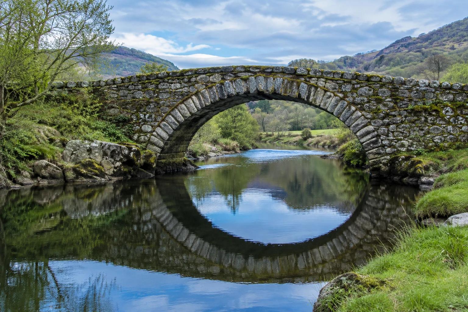 An old stone arch bridge crosses a calm river in a green valley, with the bridge and cloudy blue sky reflected in the water in a near-circular shape.