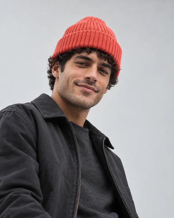 A young adult man with short dark curls wears a coral rib-knit beanie and a charcoal zip jacket over a dark crewneck, looking down toward the camera with a…