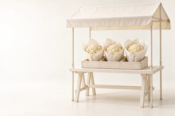 A pale wooden market stall with an off-white canvas canopy displays three cauliflower heads wrapped individually in cream paper in a shallow wooden crate…