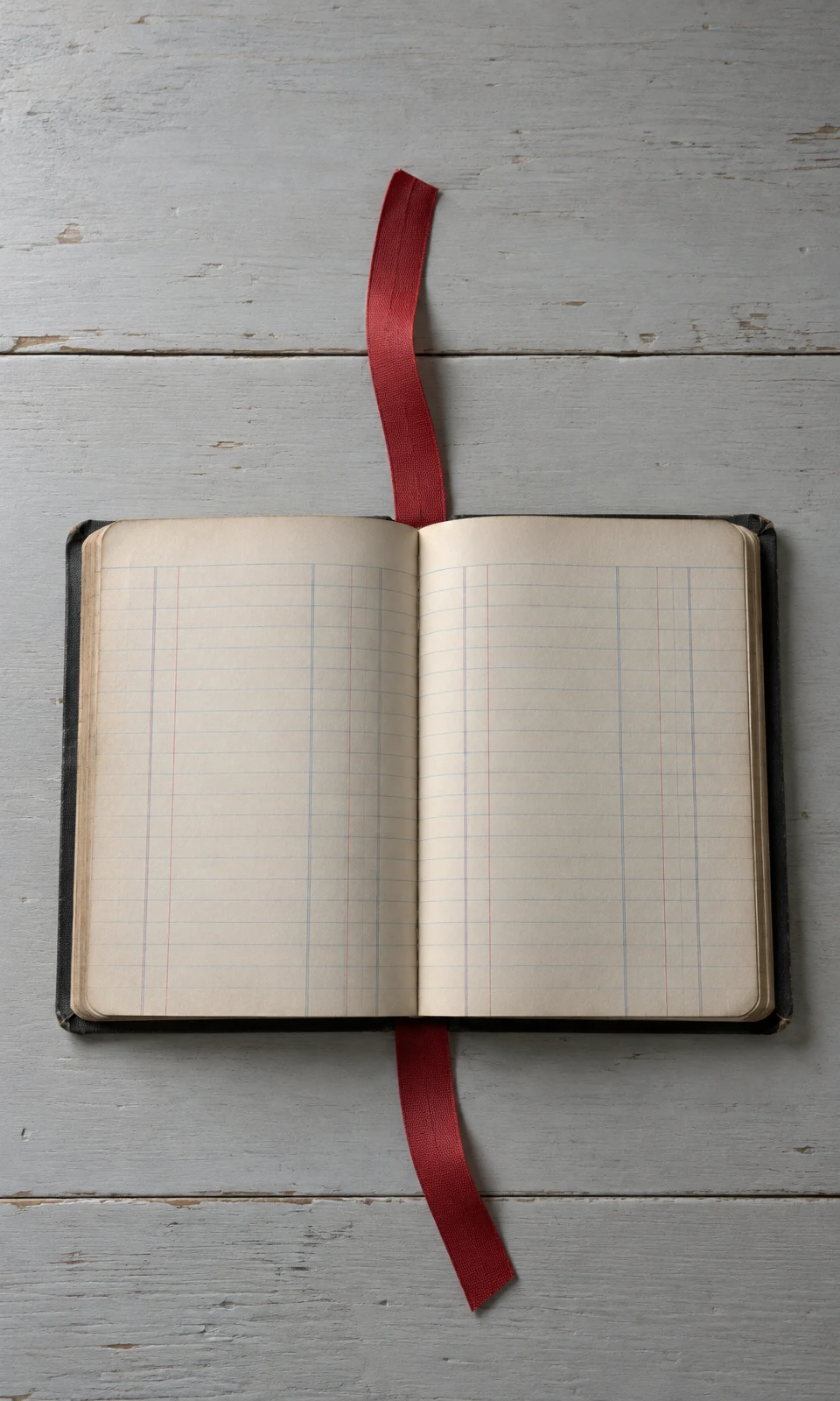 An open, gently aged charcoal-cloth ledger with blank cream pages and faint blue and red accounting rules rests on a pale gray painted wooden table.