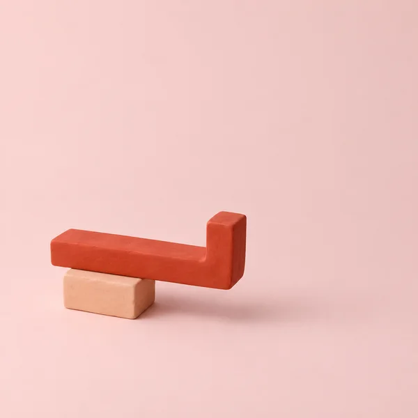 A matte vermilion bar with a blunt upward bend at its right end rests across a pale-peach rectangular clay block against a seamless chalk-pink background.