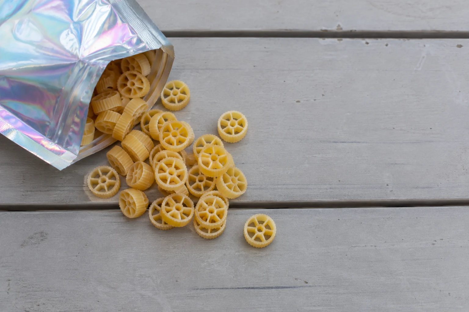 An iridescent foil pouch spills golden wagon-wheel pasta onto a pale grey, weathered plank tabletop, with open space across the right side.