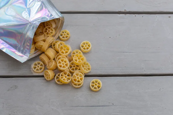An iridescent foil pouch spills golden wagon-wheel pasta onto a pale grey, weathered plank tabletop, with open space across the right side.
