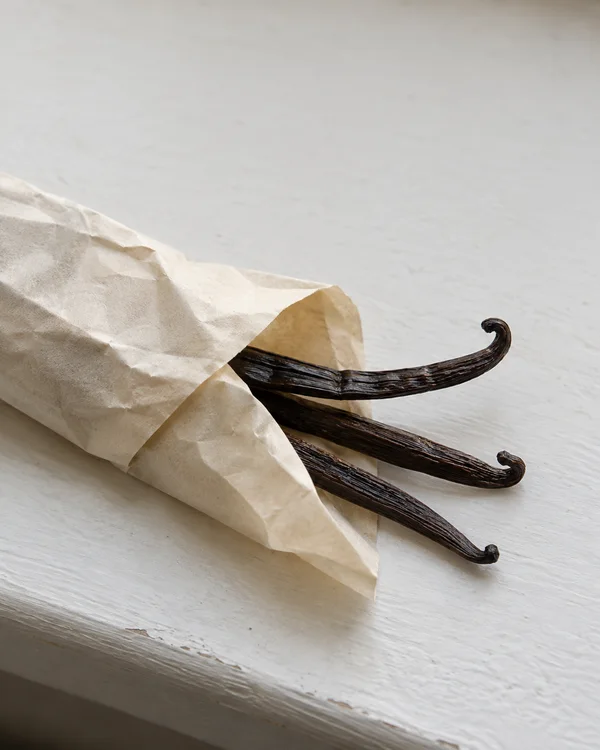 Three dark-brown vanilla pods extend from a crumpled cream-paper wrapper on a pale painted windowsill, with ample empty space above and a narrow strip of the…