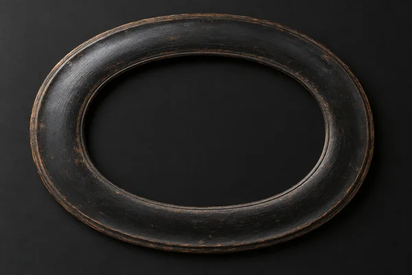 An aged, dark oval wooden picture frame lies horizontally on a charcoal-black surface, with a large empty center and warm brown wear along its rim.
