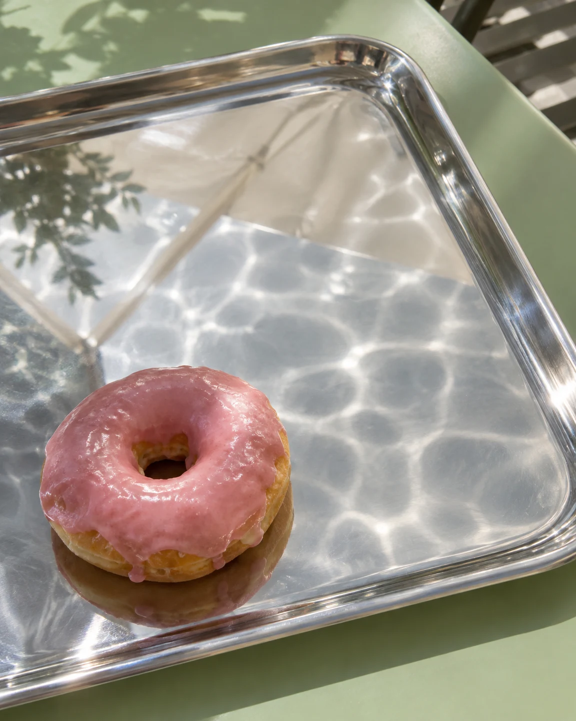 A pink-glazed ring doughnut rests near the lower-left of a polished metal tray on a pale-green table.
