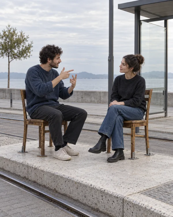Two adults sit facing one another on worn wooden chairs atop a raised concrete tram platform, with one gesturing as the other watches; rails, a shelter, water…