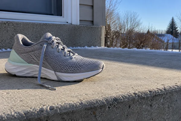 A grey mesh running shoe with a pale mint heel insert and loose laces rests on a sunlit concrete step, with snow along the rear edge and a winter landscape…