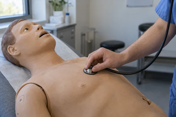 A trainee in a blue scrub sleeve presses a black stethoscope against the chest of a tan teaching mannequin on an examination couch in a clinical skills room.