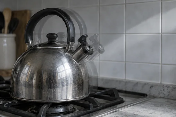 A reflective stainless-steel kettle with a black arched handle sits on a gas stovetop against pale square tiles.