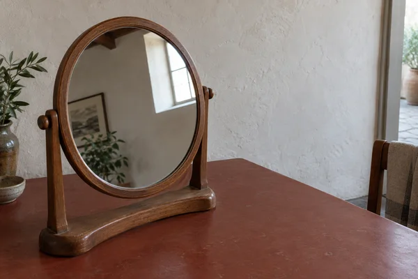 A round wooden swivel dressing mirror stands on a brick-red tabletop against a pale plaster wall, reflecting a bright window, framed picture and greenery; a…